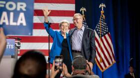Elizabeth Warren smiles and waves while standing on stage in front of an American Flag backdrop. Her partner has his arm wrapped around her.