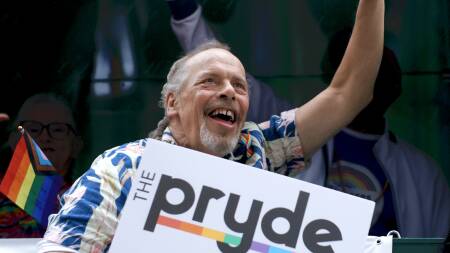 A man in a Hawaiian shirt holds up a sign that says "The Pryde" with a "y." In the background is a small "progressive Pride flag," which includes strips for LGB people, transgender and nonbinary people, Black and brown people, and people with HIV/AIDS.