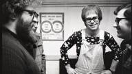 A black and white photo of Elton John backstage in 1970, wearing white overalls and a dark-colored long-sleeved shirt with stars.