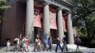 About 20 people walk along a sidewalk outside a large building with columns and hanging red flags with the Harvard seal.