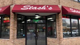 A restaurant front with red awnings reads “Stash’s.” Neon signs say "Pizza," "BBQ," "Cheesesteaks."