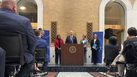 A man in a suit stands at a podium, speaking to a crowd in a hall at the State House.