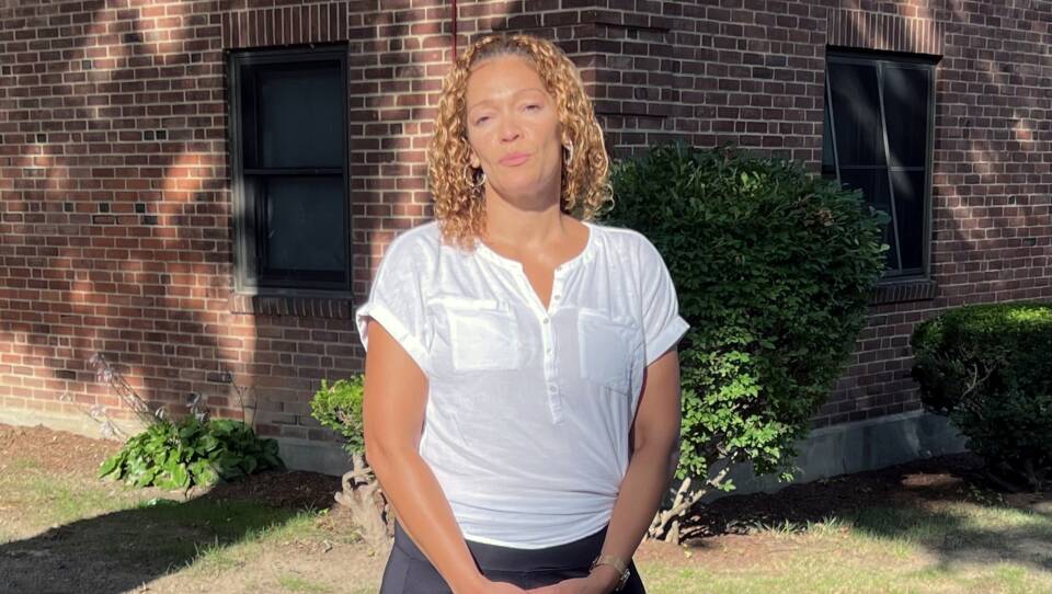 A woman holds her hands in front of her as she stands on the grass outside a brick building.