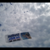 A banner planes, towing a banner ad soars in the sky, which is blanketed with clouds with patches of blue poking through