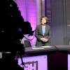 The governor stands on GBH's purple set in a radio headset and blazer, listening intently. She's facing a camera that stands in the foreground, obscured in the shadows.