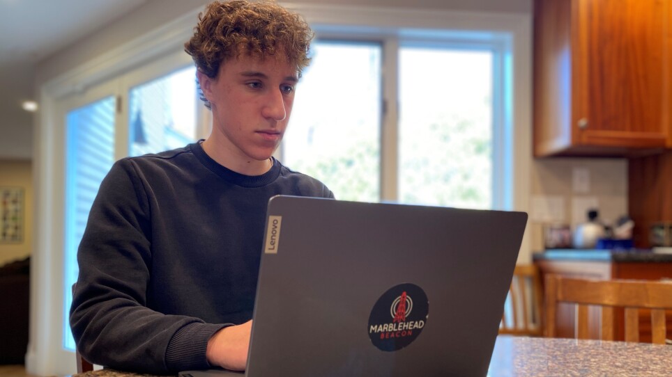 A young man sits at a kitchen counter on his laptop, which has a sticker with the words "Marblehead Beacon" on it.