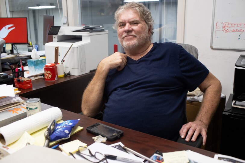 A man in a blue T-shirt with graying hair poses for the camera, sitting behind his cluttered desk