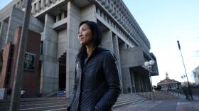 A woman with her hands in the pockets of her black jacket stands on the sidewalk outside a large concrete building.