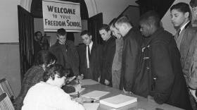 Young men of different races stand looking over a table as two women write on slips of paper.