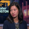 A woman in a blazer speaks on a TV show with the Greater Boston logo in the background