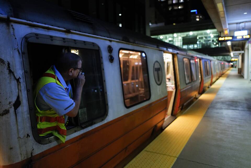 A subway train at night, with a conductor leaning out of a window speaking into an intercom.