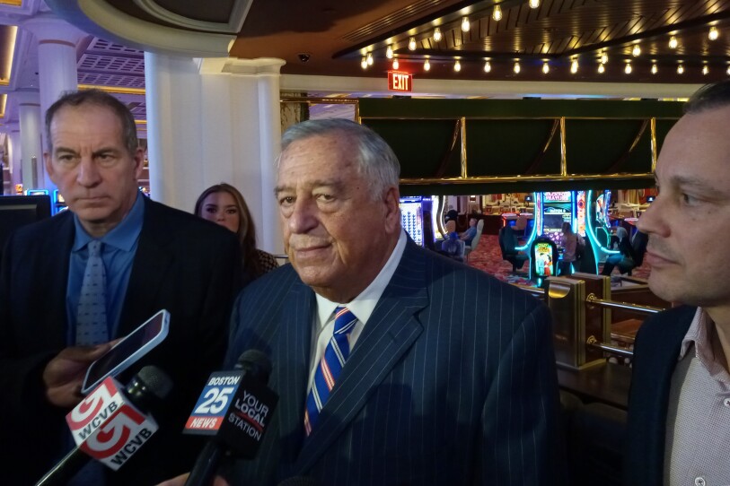 A man in a striped suit fields questions from reporters, who are holding up phones and microphones to him. In the background, the bright neon of games and gambling machines shines out.