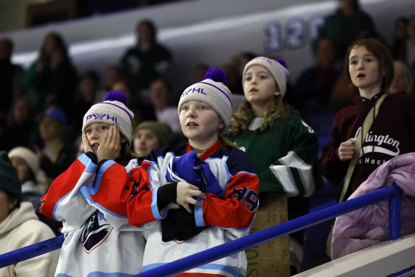 Young girls in hockey jerseys stand in an arena and watch intensely. One of them had hands on her face.