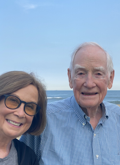 A man and woman smile at the camera against blue sky and the ocean in the background