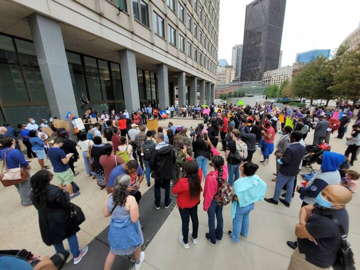 A group of 100 protestors stand outside the federal building, many holding colorful posters