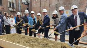 A group of men and women in dress clothes and hard hats fling dirt into the air with shovels.