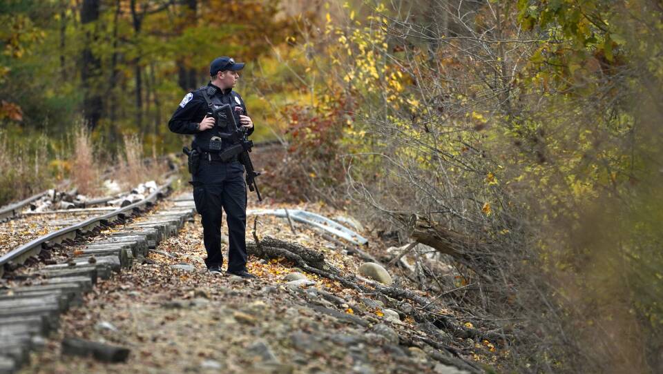 A police officer carrying a gun stands alongside a railroad track amid fall foliage
