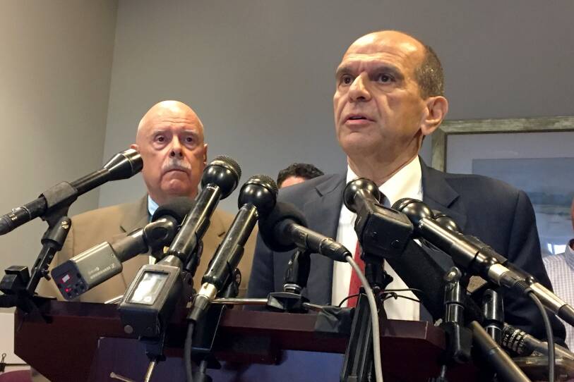 Two men in suits stand at a podium, looking somber. One is speaking into nearly a dozen microphones that are attached to the podium.