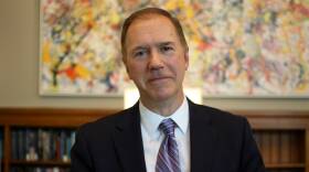 A man in a suit and tie stands in front of an abstract painting and bookshelves.
