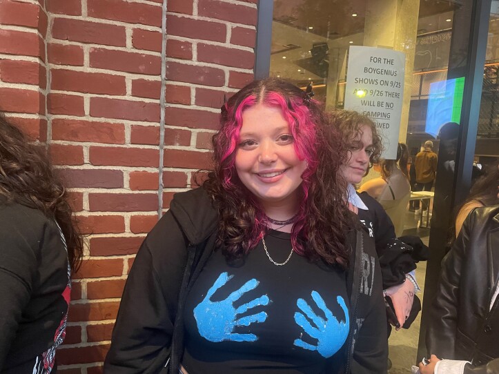 A young woman standing in line for the boygenius concert. She has wavy shoulder-length hair. The from of her hair is colored a bright magenta. She is wearing a black sweatshirt, that has two blue hands on it. The effect --or visual gag--is as if someone whose hands had just been dipped in blue paint has touched her breasts.