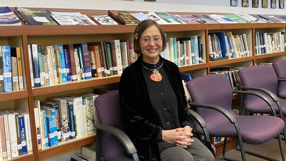 A woman in a black button-down shirt, velvet jacket, and a silver-and-orange necklace sits in front of a stack of books, smiling.