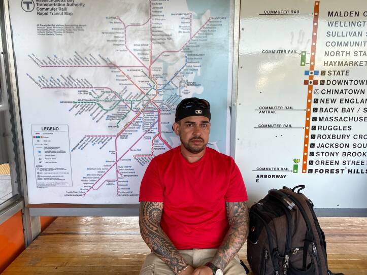 A man in red T-shirt wearing a backwards baseball cap and sunglasses on top of his head sits on a wooden bench, leaning against a map of the MBTA system.