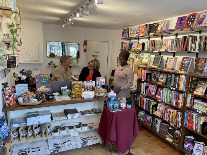 Three women gather around the check out counter at a small bookstore. To the right of the counter is a wall lined with bookshelves. The front of the is a display of merchandise such as book bags and notebooks