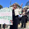 A woman with blond hair and glasses speaks into a microphones. Standing around her are people with signs showing support for funding programs for domestic violence survivors.