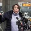 A woman with short brown hair is mid-speech in front of a cluster of microphones, wearing a light purple turtleneck under a winter coat with the reflection of an MBTA bus in the background.