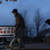 A man outdoors at twilight, leaning down to collect a sign reading vote here with an arrow pointing to the right.