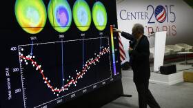 A man holds a microphone and gestures to part of a roughly 10-foot tall display showing a timeline and illustrations of the Earth's ozone layer over the decades.