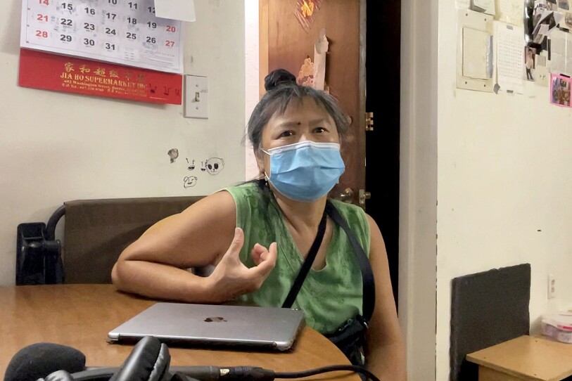 A woman wearing a mask sits at a small wooden table in front of a laptop