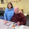 A woman stands with her arm around her adult son. Markers and a coloring book are on a table in front of them.