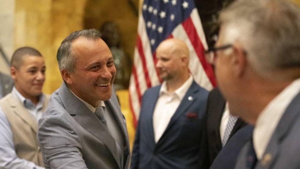 A smiling man wearing a suite reaches over to shake hands with another man.