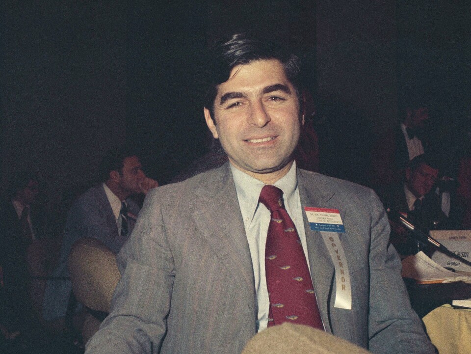 A man in a suit and tie smiles for the camera. He's wearing a name tag with a ribbon that reads 