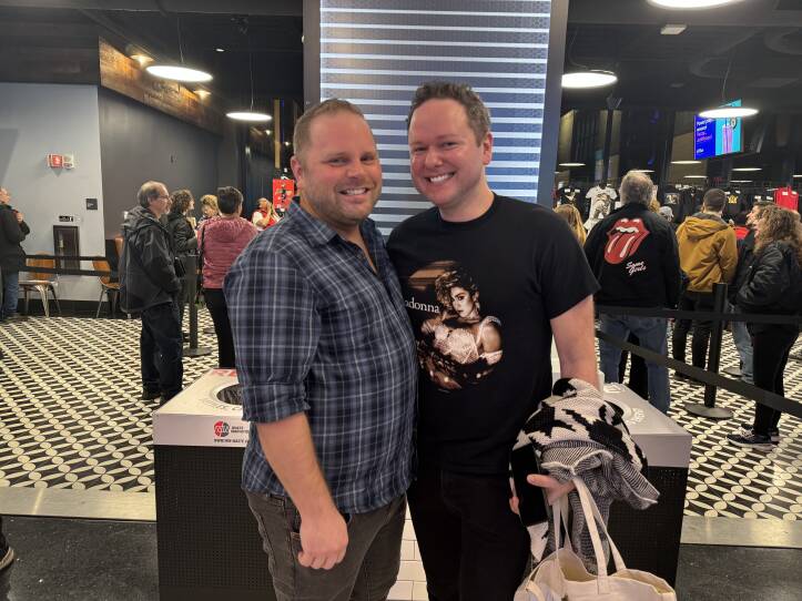 Two men stand together, one wearing a blue plaid shirt and the other a shirt with a photo of pop singer Madonna on it.