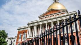 The sun shines bright on the golden dome of the Massachusetts State House, which stands behind an iron fence.