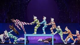Performers in matching multicolored onesies balance across a table, holding each other up