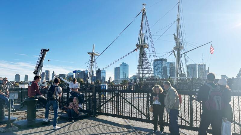 Two people stand behind a metal fence as they look at an old wood-masted sailing ship.