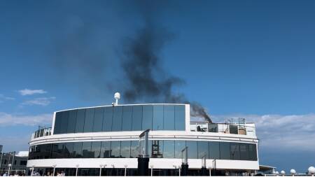 Black smoke rises above a large cruise ship and into the blue sky.