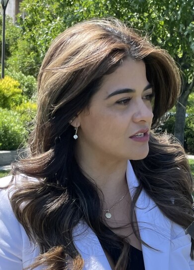 A woman with long, wavy brown hair faces to the side as she speaks with someone outside on a sunny day.