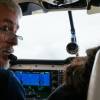 A pilot, with shortly cropped white hair and glasses, turns to his left. Large pilot headphones are over his ears, and a microphone is over his mouth. He sits in the cockpit of a plane next to a passenger with long dark hair.