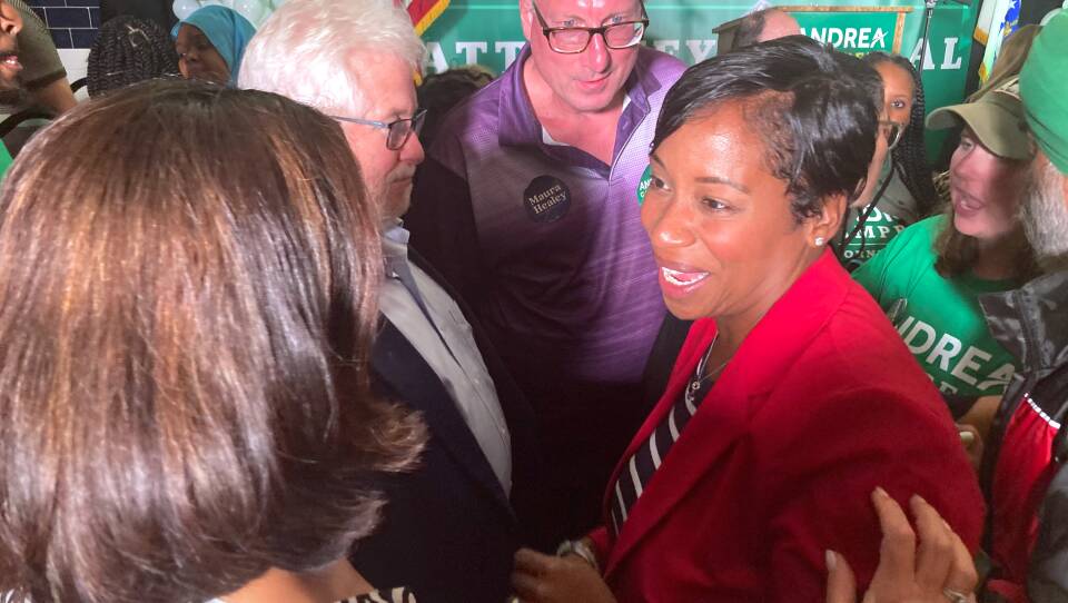 Andrea Campbell, in a red jacket, stands in a crowd of people in front of a green campaign sign.