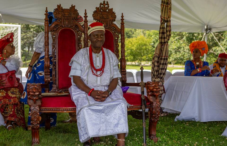 A man wearing a white outfit with a red cap and red beaded necklace sitting on a large red chain with hands clasped in his lap.
