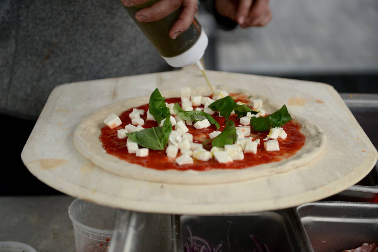 A close-up on a small pizza pie, with sauce, cheese and basil, with a hand squeezing olive oil out of a bottle in circles around the top.