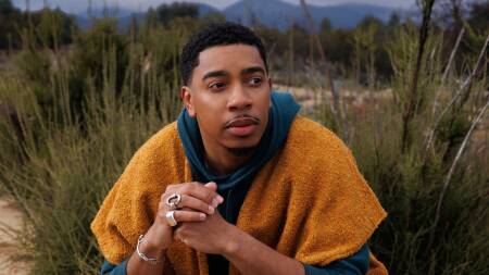 Christian Sands crouches in a clump of beach grass looking off into the distance.