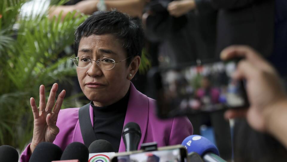A woman in a pink jacket speaks before a group of reporter with microphones and cellphones.