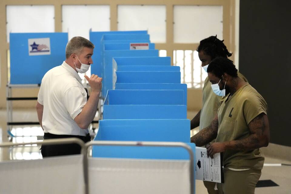 A corrections officer in uniform speaks two inmates wearing olive drab clothing as they stand at a row of blue voting booths.