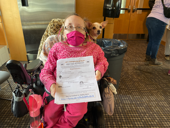 A woman in a wheelchair holds up a paper that says 'Part-time PCA needed.' A small chihuahua sits behind her.