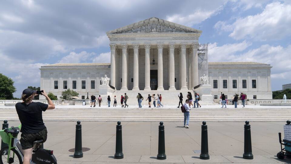 A couple dozen people on foot and on bikes stand outside the Supreme Court building, some taking photographs with their phones.
